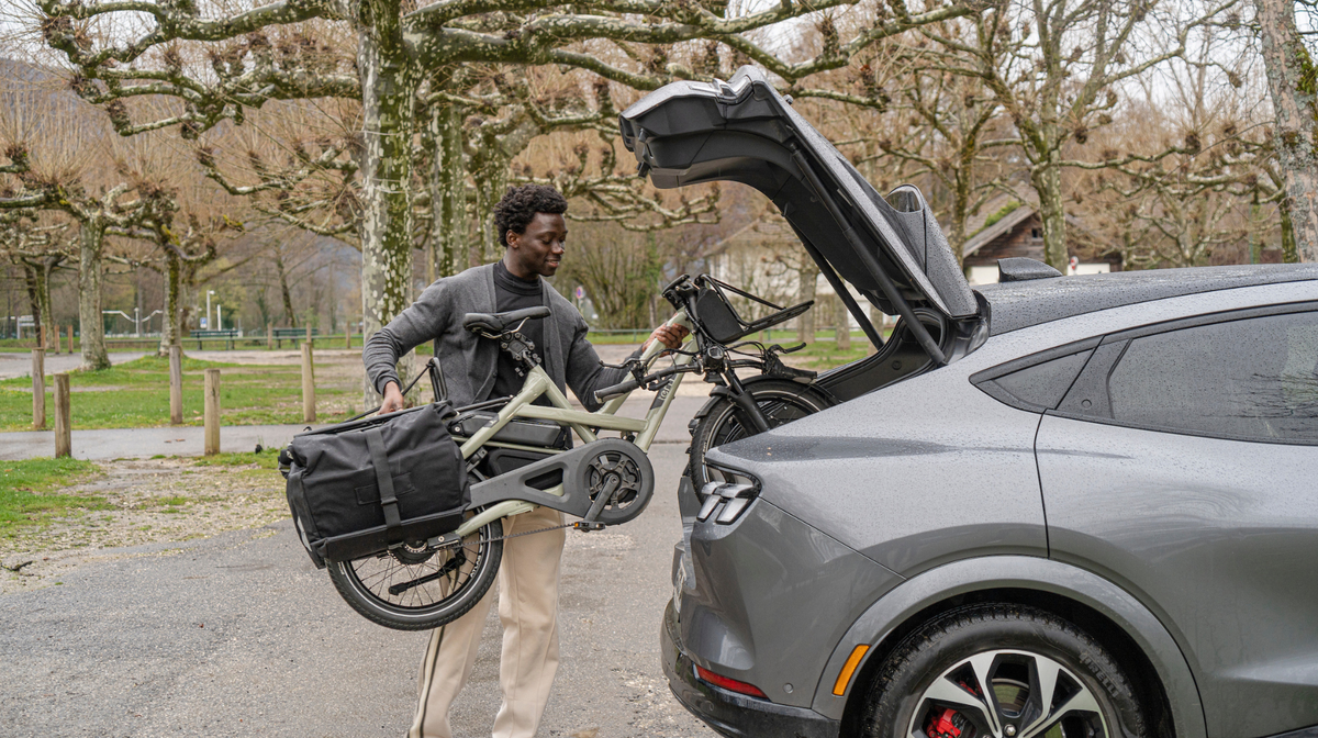 A man loading a folding Tern electric bicycle into the open trunk of a gray SUV in an outdoor park setting.