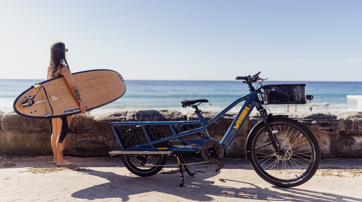 A blue Yuba Spicy Curry cargo bicycle parked on a beach next to a woman holding a wooden surfboard, with the ocean and sky in the background.