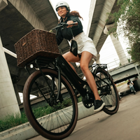 A woman wearing a white helmet rides a black Lekker Jordaan step through electric bicycle with a brown wicker basket under a concrete bridge. She is wearing a black jacket, a white skirt, and black sneakers.