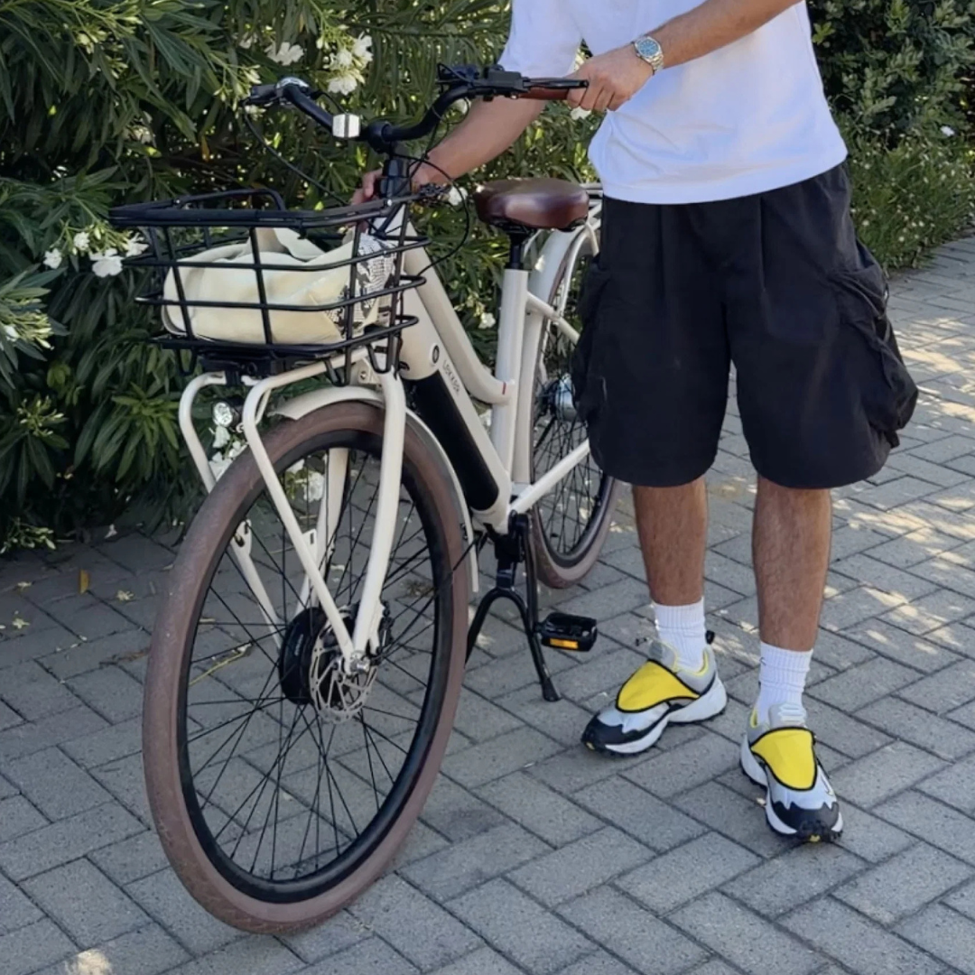 A person standing outdoors on a paved path next to a Linen Cream Lekker Jordaan step-through electric bicycle with a front basket, wearing black cargo shorts, a white t-shirt, and yellow-and-black sneakers.