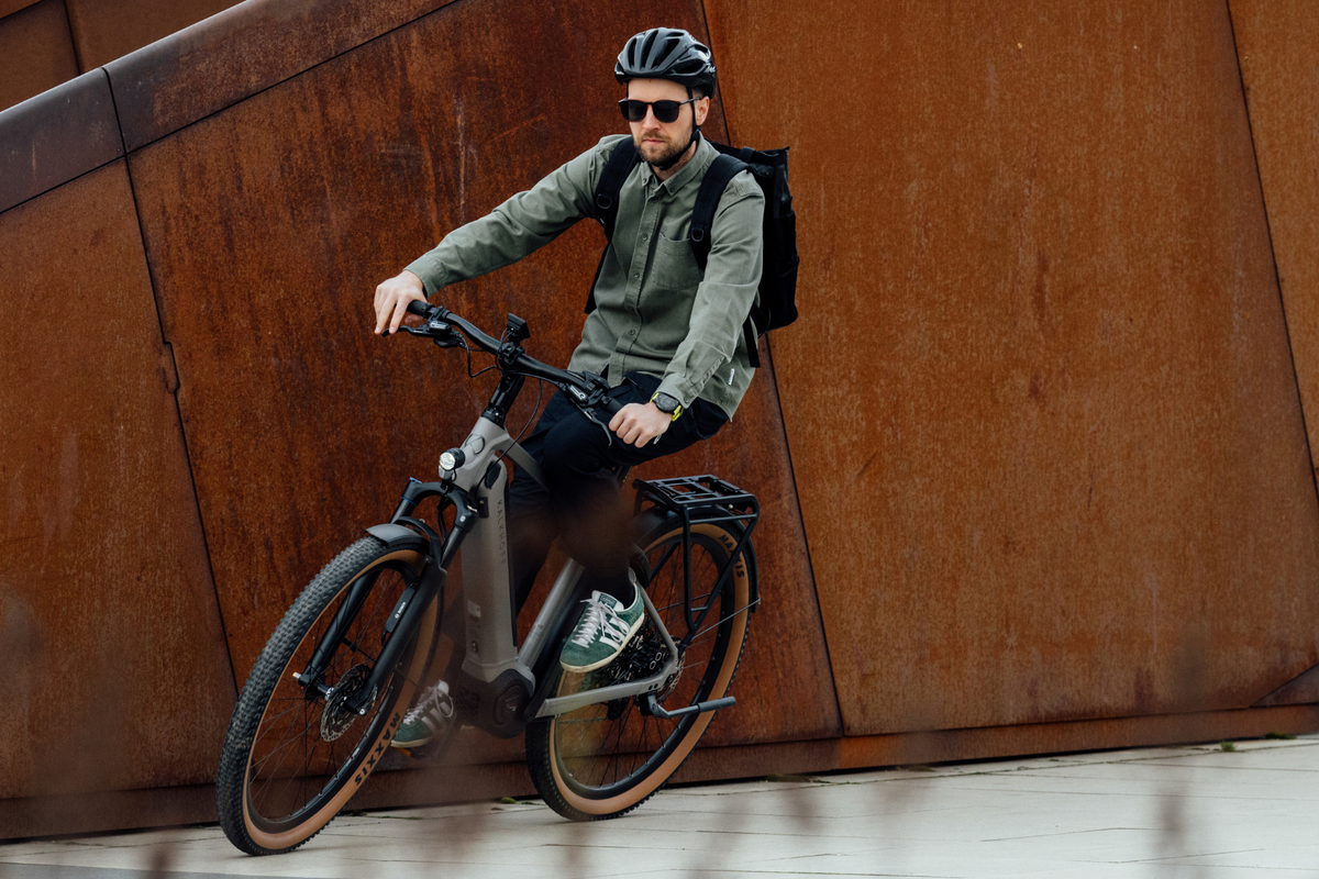 A man wearing a black bicycle helmet, sunglasses, and a green jacket rides a grey Kalkhoff electric bicycle against a large rusted metal wall.