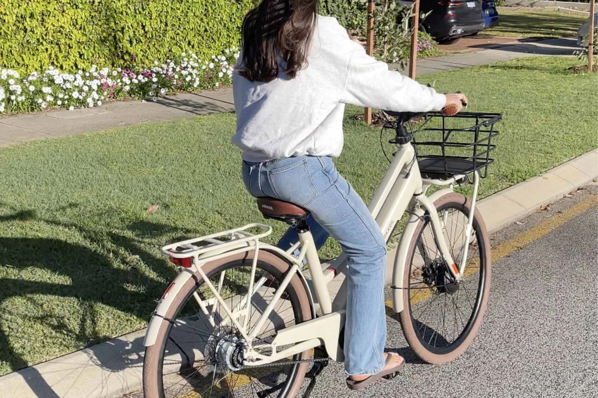 A woman wearing a white sweatshirt and blue jeans rides a linen cream Lekker Jordaan step-through electric bicycle with a front basket and rear rack on a paved path next to a grassy lawn.
