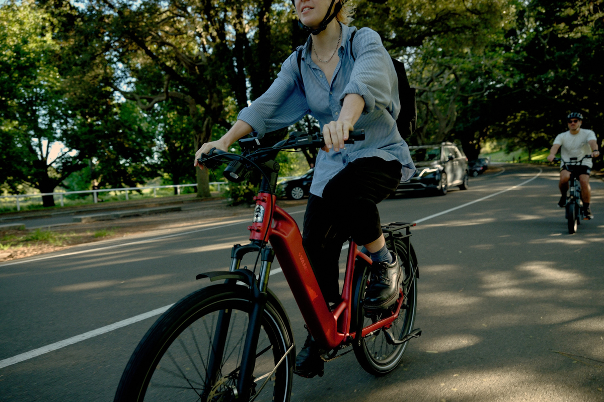 A woman riding a red Vamos Rapido electric bicycle on a tree-lined road, wearing a light blue button-up shirt, black pants, and a black helmet. Another cyclist is visible in the background.